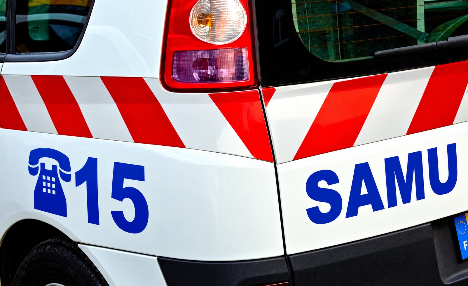 Vue rapprochée de l'arrière d'une ambulance blanche du SAMU, avec le numéro 15, un téléphone bleu et des bandes rouges réfléchissantes.