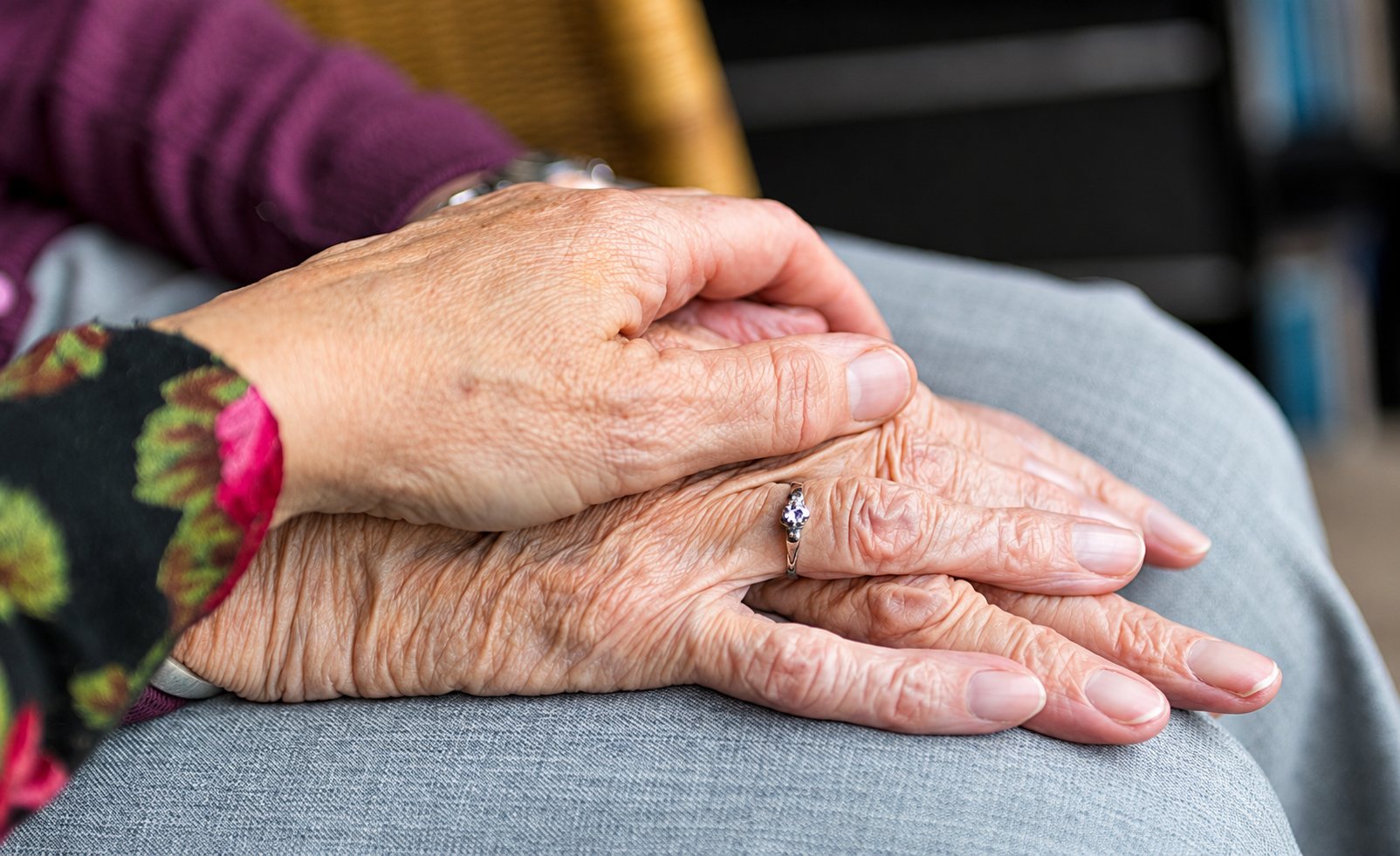Une main se pose avec douceur sur les mains ridées d'une personne âgée portant une bague, posées sur ses genoux.