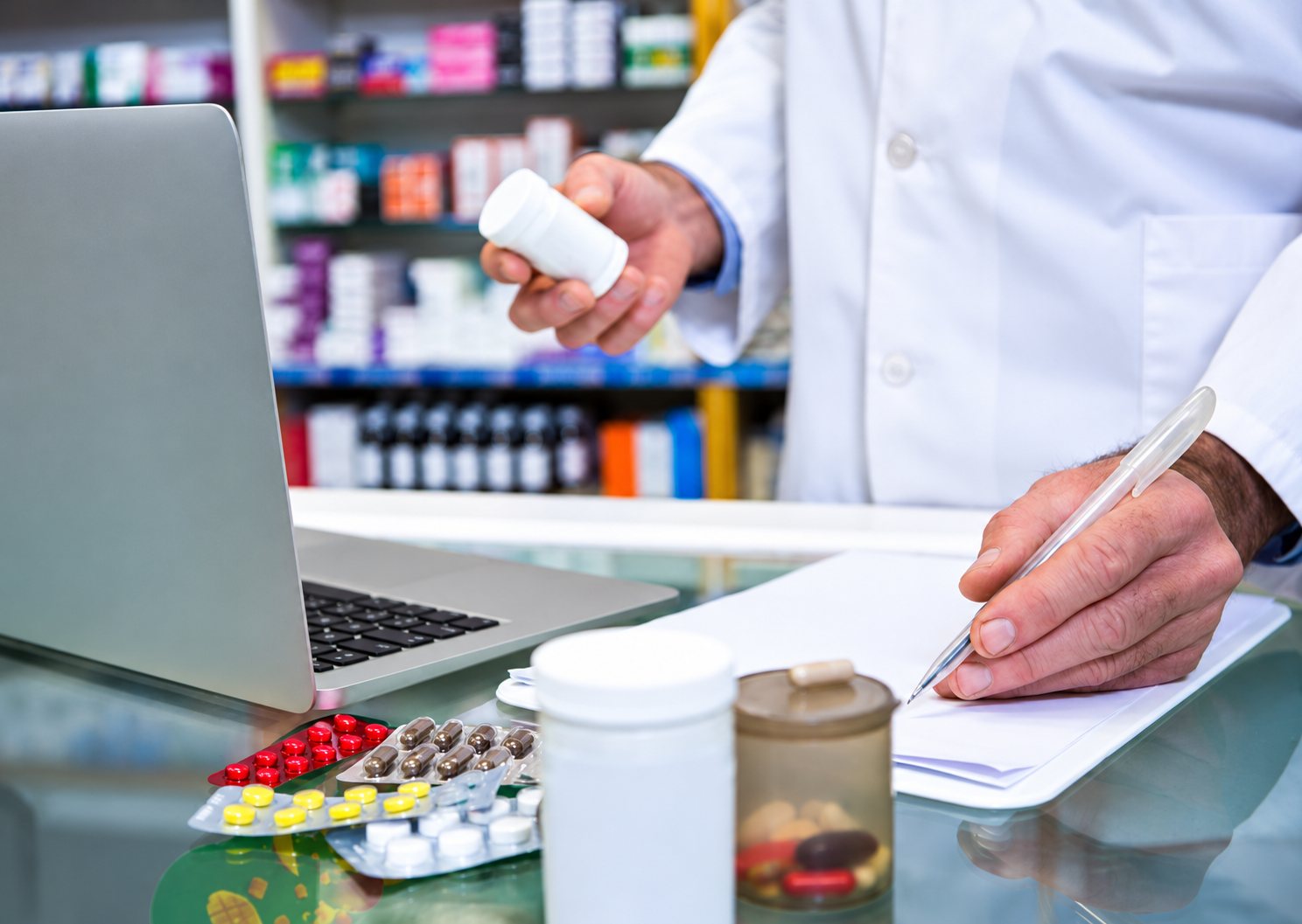 Un pharmacien en blouse blanche tient un flacon de médicaments et écrit sur un papier au comptoir, près d'un ordinateur portable et de plaquettes de comprimés.