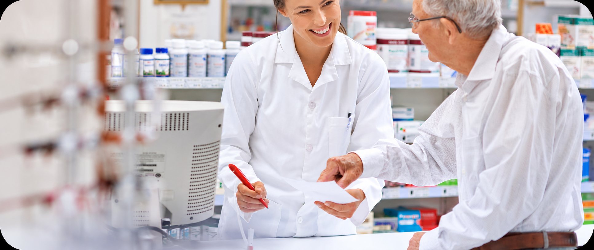 Une pharmacienne en blouse blanche sourit en montrant une ordonnance à un client âgé au comptoir, devant des étagères de médicaments.