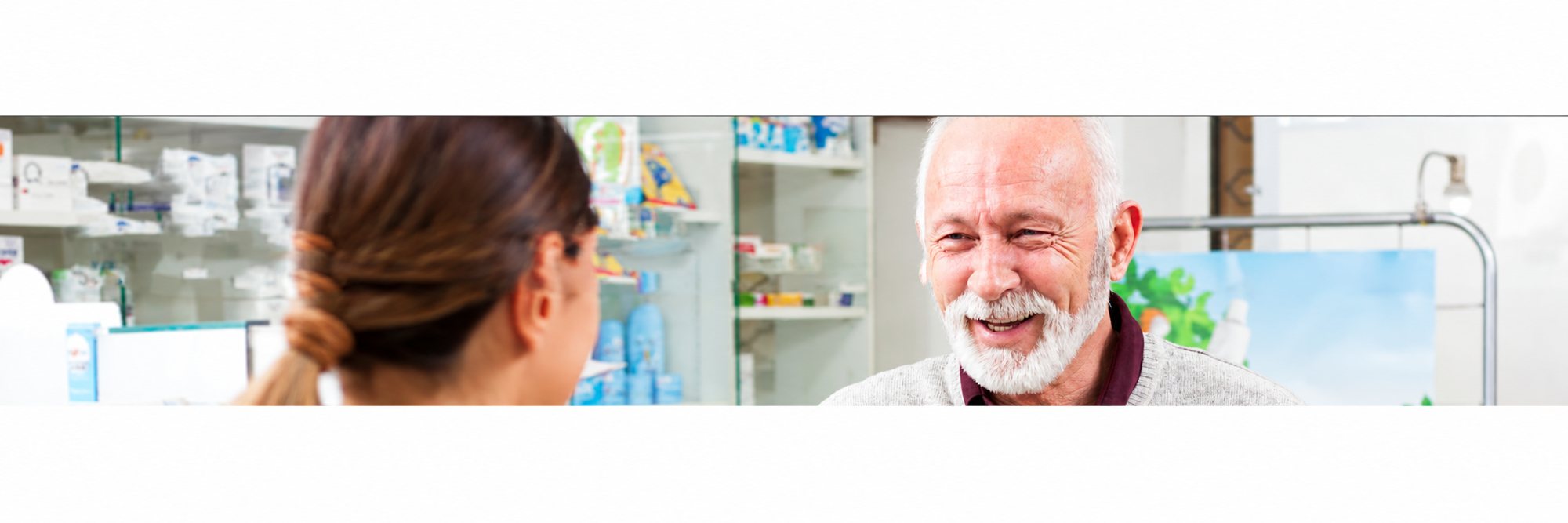 Une femme vue de dos discute avec un homme âgé à barbe blanche qui sourit, dans une pharmacie aux étagères de produits floutées.