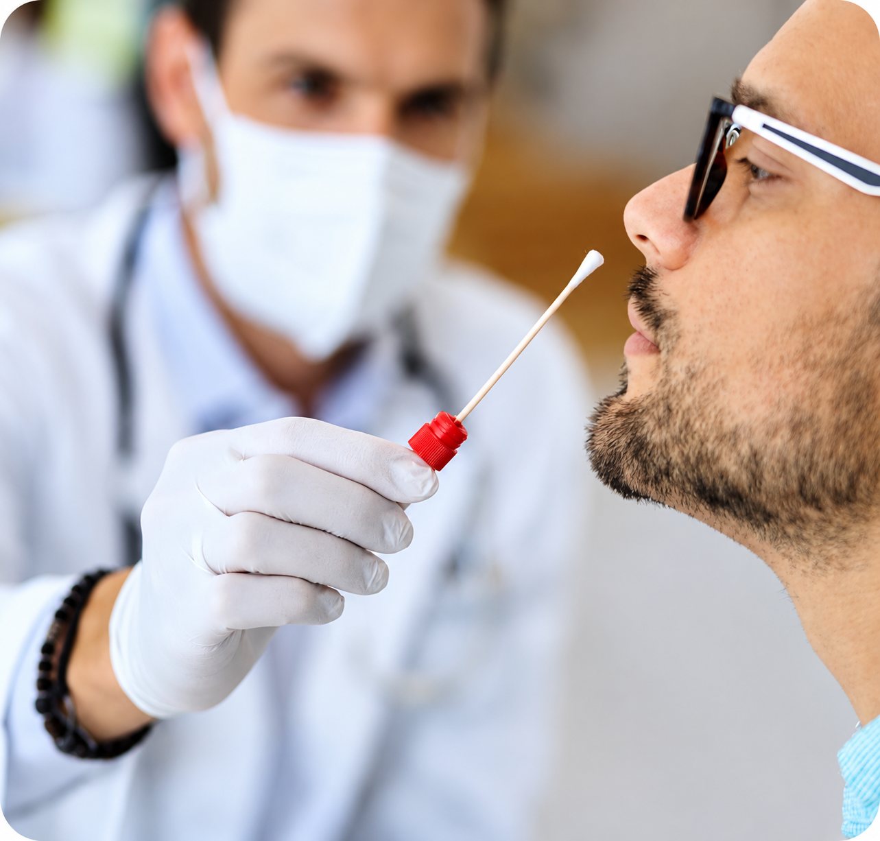 Un soignant masqué et ganté approche un écouvillon à embout rouge du nez d'un patient barbu portant des lunettes.